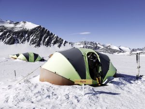 A person in winter gear stands at the entrance of a green tent labeled Byrd on the snowy landscape of Antarctica, with other tents and snow-covered mountains in the background under a clear blue sky—a scene supported by Antarctic Logistics and Expeditions.
