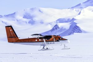 A red and white ski plane is landing or taking off on a snowy, icy surface in Antarctica, with snow-covered mountains in the background under a clear blue sky—perfect for Antarctic Logistics and Expeditions.