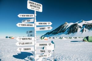 A signpost at Union Glacier, Antarctica, managed by Antarctic Logistics and Expeditions, shows distances to major cities worldwide. Snow, mountains, and tents are visible in the background under a clear blue sky near the South Pole.