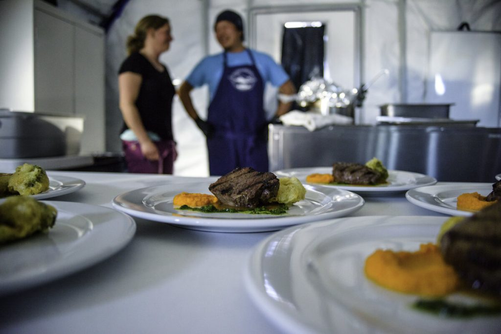 Plated gourmet meals with meat and colorful purees are arranged on a table in the foreground, while two people—perhaps discussing Antarctic Logistics and Expeditions—converse in a kitchen setting in the blurred background.