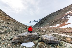 A person in a red jacket sits on a large rock in a rocky, snow-dusted Antarctic valley, with another hiker ahead and snowy peaks in the distance—capturing the spirit of adventure found on Antarctica expeditions.