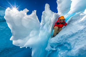 A person in a red jacket and orange helmet smiles while crouching inside a jagged blue ice crevasse in Antarctica, with bright sunlight streaming through the icy formations against a clear South Pole sky.