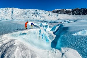 Two people in bright jackets stand on the edge of a deep crevasse in a vast, icy Antarctic glacier landscape under a clear blue sky, with snow-covered mountains in the background—capturing the spirit of Antarctic Logistics and Expeditions.