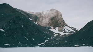 A rugged, snow-dusted mountain peak rises between dark rocky slopes under a cloudy sky in Antarctica, with patches of snow scattered across the terrain and a flat, icy surface—a scene typical of Antarctic Logistics and Expeditions.