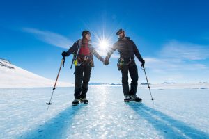 Two people in winter gear hold hands on a sunlit Antarctic landscape. Each holds walking poles as the South Pole sun shines brightly between them, casting long shadows across the icy ground.