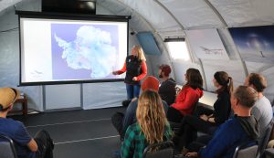 A woman stands by a projector screen displaying a map of Antarctica, giving a presentation on Antarctic Logistics and Expeditions to a seated group of people in a tent-like room decorated with South Pole photos.