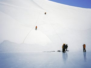 Several people wearing bright jackets are on a snowy, icy slope in Antarctica. Two climb the steep incline with ropes, while others stand below observing and photographing, highlighting an adventure supported by Antarctic Logistics and Expeditions.