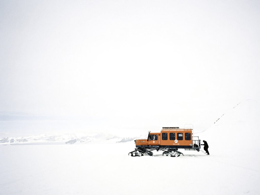 A person stands next to an orange snowcat vehicle on a vast, snowy landscape in Antarctica with distant mountains under an overcast sky, capturing the spirit of Antarctic Logistics and Expeditions.