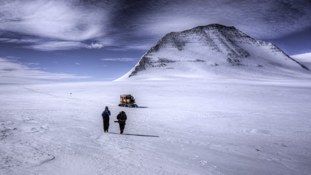 Two people in winter gear trek across a snowy, icy landscape in Antarctica toward a rugged, snow-covered mountain, with a bright orange Antarctic Logistics and Expeditions vehicle parked in the distance under a partly cloudy sky.
