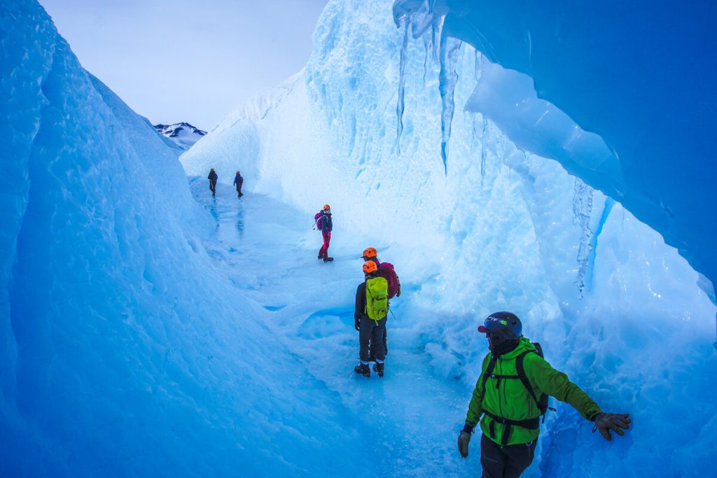 A group of people in winter gear and helmets walk and explore a narrow, icy blue glacier tunnel surrounded by tall frozen walls under a pale sky, experiencing the wonders of Antarctica with Antarctic Logistics and Expeditions.