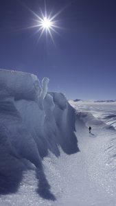 A person stands near the base of a tall, jagged blue ice wall under a bright sun in Antarctica's vast snowy landscape, with clear blue sky—an awe-inspiring scene made possible by Antarctic Logistics and Expeditions.