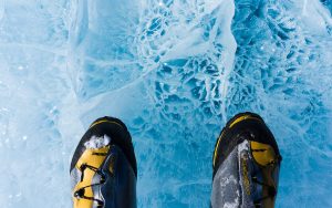 A close-up view of two boots standing on textured, cracked blue ice in Antarctica, with snow on the boots and intricate ice patterns visible beneath.