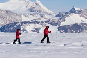 Two people in red jackets cross a snowy, icy landscape on skis in Antarctica, with snow-covered mountains and a clear blue sky in the background—an adventure made possible by Antarctic Logistics and Expeditions.