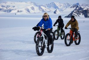 Three people ride fat-tire bikes on snow near the South Pole, dressed in winter jackets, gloves, and sunglasses, with snowy mountains and a clear sky in the background.