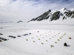 A large camp with numerous tents is set up on a snowy landscape near snow-covered mountains under a partly cloudy sky. Several structures and vehicles are scattered throughout the camp, typical of Antarctic Logistics and Expeditions in Antarctica.