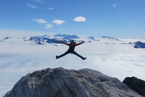 A person in winter gear jumps with arms and legs outstretched on a rocky peak, with snowy mountains and Antarctica’s vast icy landscape in the background under a clear blue sky—an epic moment courtesy of Antarctic Logistics and Expeditions.