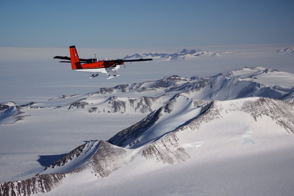 A red twin-engine plane with skis from Antarctic Logistics and Expeditions soars above Antarctica's snow-covered mountains and icy terrain under a clear blue sky.