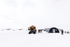 A snowy landscape in Antarctica with an orange snow vehicle, several people near a blue tent, and two small red airplanes in the background. Bicycles and equipment from Antarctic Logistics and Expeditions are visible on the icy ground.