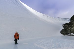A person in a red parka stands on a frozen, icy surface beneath a massive, curved snow-covered slope with rocky terrain on one side under a cloudy South Pole sky in Antarctica.