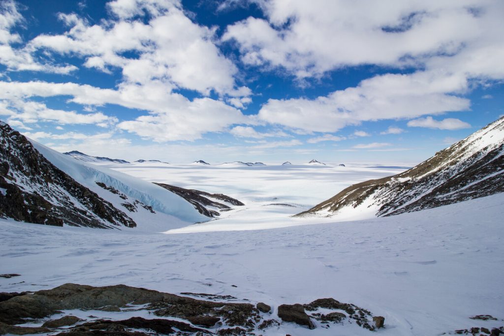 Snowy mountains frame a wide, icy Antarctic valley under a partly cloudy blue sky, with distant dark peaks rising above a vast, frozen expanse near the South Pole in the background.