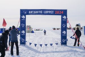 A cross-country skier approaches the finish line under a blue banner reading Antarctic Loppet 2024, surrounded by spectators and flags on a snowy landscape.
