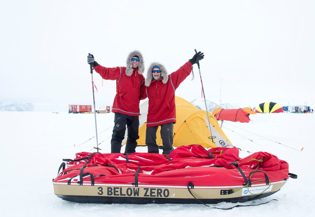 Two people wearing red parkas and sunglasses stand smiling with arms raised on a snowy landscape, behind a sled labeled 3 BELOW ZERO and in front of yellow tents and camping gear in the snow.