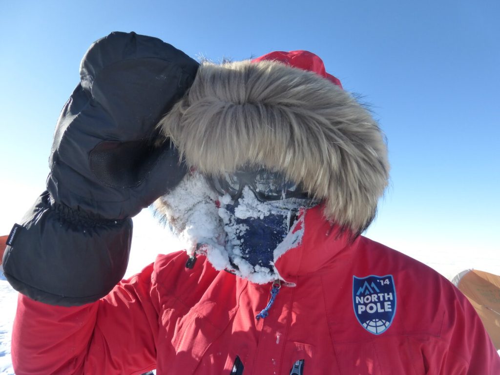A person in a red parka with a fur-lined hood and large mittens stands in a snowy landscape, their face mostly covered in ice and frost; a badge on the parka reads North Pole.
