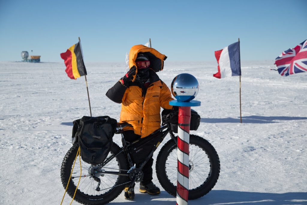 A person in a bright orange parka stands in snowy terrain with a fat-tire bike, saluting next to a striped pole topped with a reflective sphere. International flags, including Belgium, France, and the UK, are in the background.
