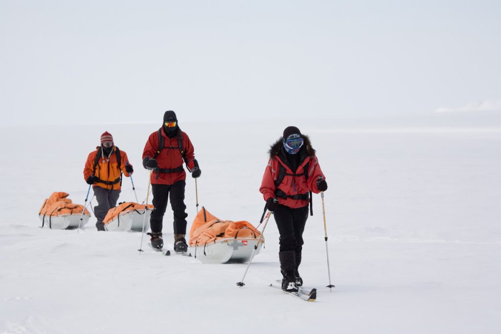 Three people wearing heavy winter gear ski across a snowy, icy landscape, each pulling a sled loaded with orange-covered supplies. The sky and surroundings are bright and white, indicating a cold, remote environment.