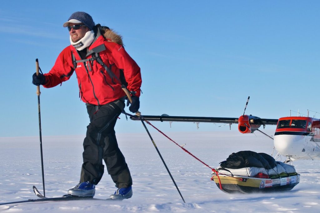 A person in winter gear skis across a snowy landscape, pulling a loaded sled. There is a small plane with skis for landing gear in the background, parked on the snow under a clear blue sky.