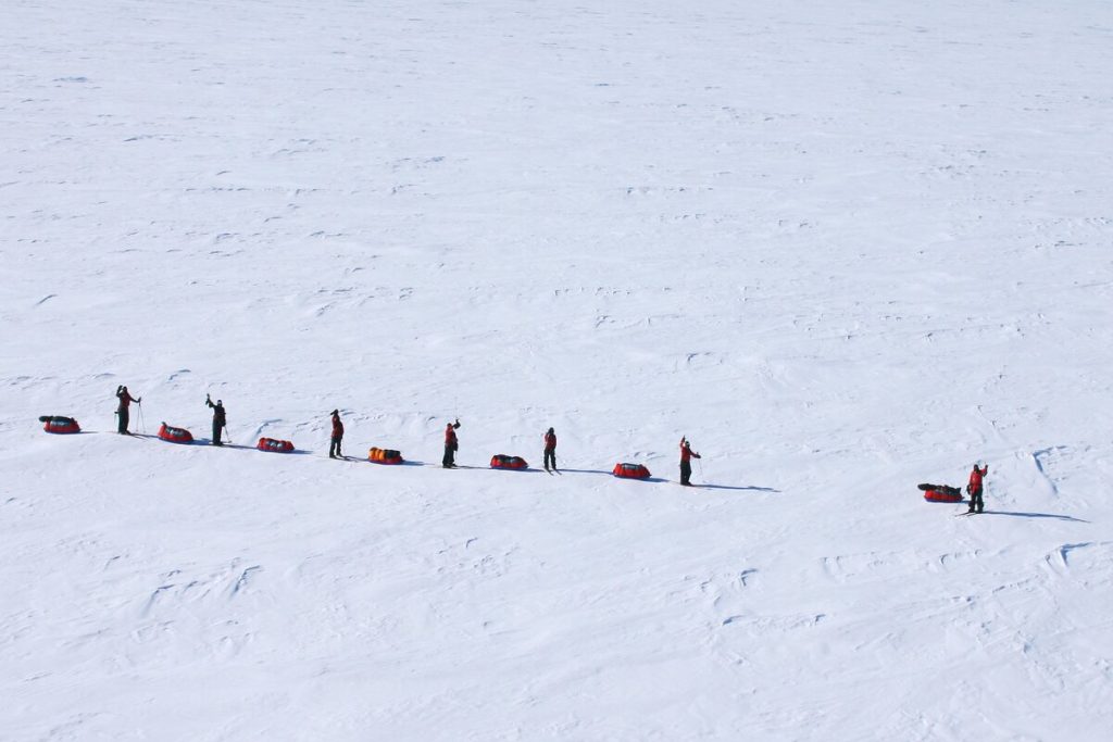 A line of eight people in winter gear pull sleds across a vast, snowy, and flat landscape under clear skies, casting long shadows on the white surface.