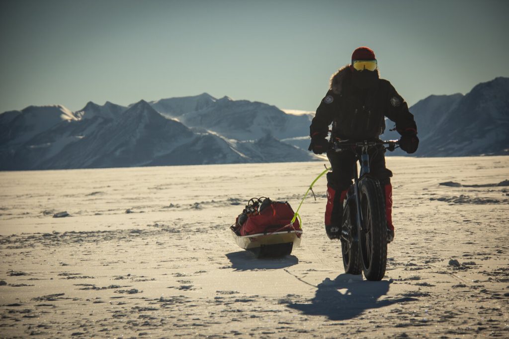 A person in cold-weather gear rides a fat-tire bike across a snowy landscape, pulling a supply sled. Snow-covered mountains are visible in the background under a clear sky.