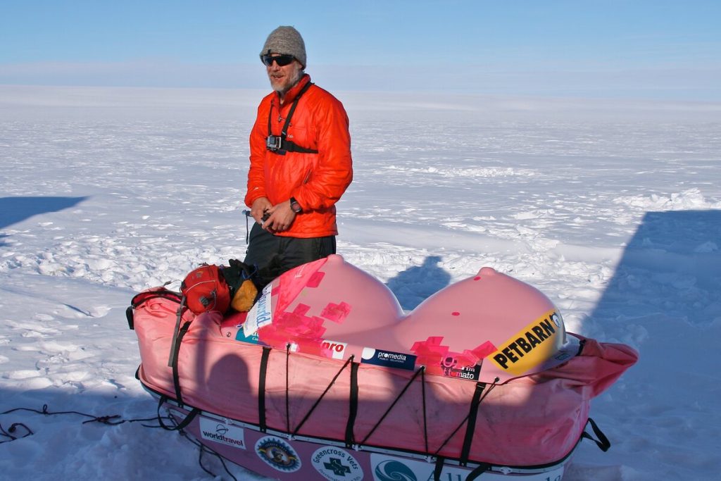 A man in an orange jacket and gray beanie stands on snow next to a pink sled loaded with gear and covered in sponsor logos, with a vast, icy landscape and clear blue sky in the background.