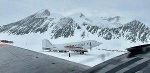 A white propeller plane is parked on snow with supplies and yellow tents nearby, ready for an overnight trip at the South Pole. Steep, snow-covered mountains rise in the background beneath a cloudy sky.