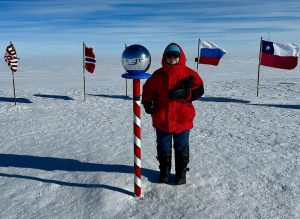 A person in a red parka stands on snow next to a striped pole with a reflective sphere, surrounded by international flags under a clear blue sky during an overnight trip at the South Pole.