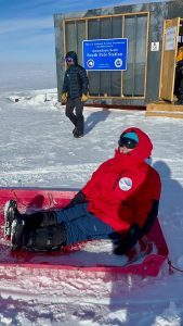 A person in a red parka and snow boots sits in a sled on the snow near the Amundsen-Scott South Pole Station, as another in winter gear stands nearby—capturing a moment from an unforgettable overnight trip at the South Pole.