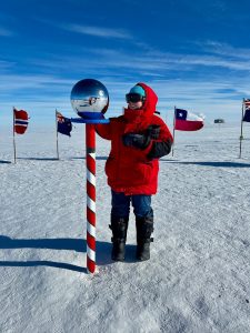 A person in a red parka and cold-weather gear stands next to a striped pole topped with a silver sphere, surrounded by snow and international flags under a bright blue sky during an overnight trip at the South Pole.