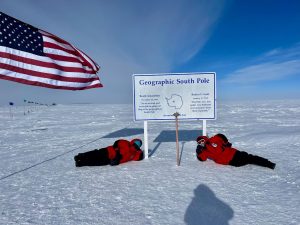 Two people in red parkas lie on snow beside a sign reading Geographic South Pole, enjoying an overnight trip at the South Pole. A large American flag flies on the left, and the icy, flat landscape stretches under a bright blue sky.