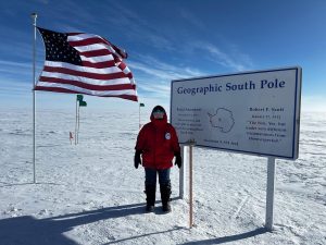 A person in a red parka stands next to a “Geographic South Pole” sign on a snowy landscape, enjoying an overnight trip at the South Pole, with an American flag and green flag behind them under a clear, sunny sky.
