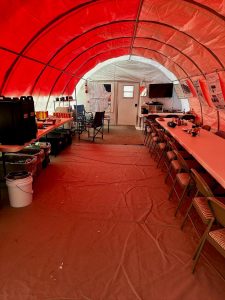 A large tent with red lighting houses long tables and chairs along both sides, set up for an overnight trip at the South Pole. Coffee dispensers, food containers, and kitchen equipment are arranged at the back, with a tarp covering the chilly floor.