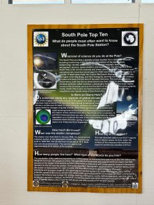 Poster titled “South Pole Top Ten” with text and images answering common questions about the South Pole Station—including science, cost, population, size, ozone hole, ice thickness, and what an overnight trip at the South Pole involves. White wall background with some reflections.