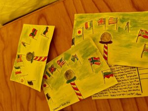 Three colorful postcards with hand-painted illustrations of various international flags on flagpoles are laid out on a wooden surface, hinting at the excitement of an overnight trip at the South Pole, one partly covering a handwritten postcard.