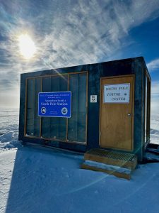 A small building labeled South Pole Visitor Center sits on snowy ground under a bright sun and blue sky at the Amundsen-Scott South Pole Station, welcoming guests arriving for an unforgettable overnight trip at the South Pole. A sign and wooden steps lead to the entrance.