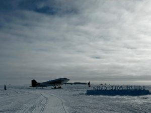 A small propeller plane sits on a snowy, icy runway under a cloudy sky. Snow blocks in the foreground spell out South Pole Camp—an inviting stop for an unforgettable overnight trip at the South Pole amid this flat, desolate landscape.