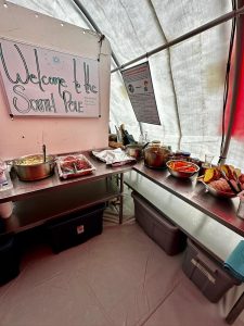 A buffet table inside a tent features trays of food, bread, and utensils under a sign reading Welcome to the South Pole—an inviting setup for an overnight trip at the South Pole, with supplies stored in bins beneath the tables.
