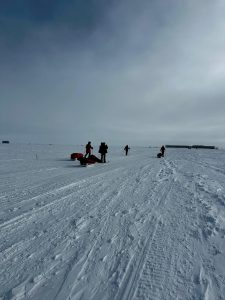 Four people in winter gear stand and kneel on a vast, snowy landscape during an overnight trip at the South Pole, pulling sleds. The overcast sky and packed snow with visible tracks lead to distant structures on the flat horizon.