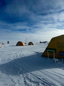 A snowy campsite with orange and yellow tents set up on a flat, snow-covered landscape under a partly cloudy sky. Tracks in the snow lead past equipment towards the tents, evoking an overnight trip at the South Pole.