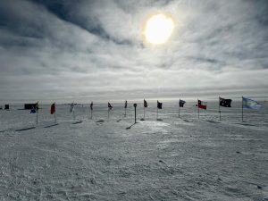Flags from several countries stand in a row on a snowy, icy landscape under a bright sun. A few small buildings in the distance hint at an overnight trip at the South Pole, making the cold and remote scene even more captivating.