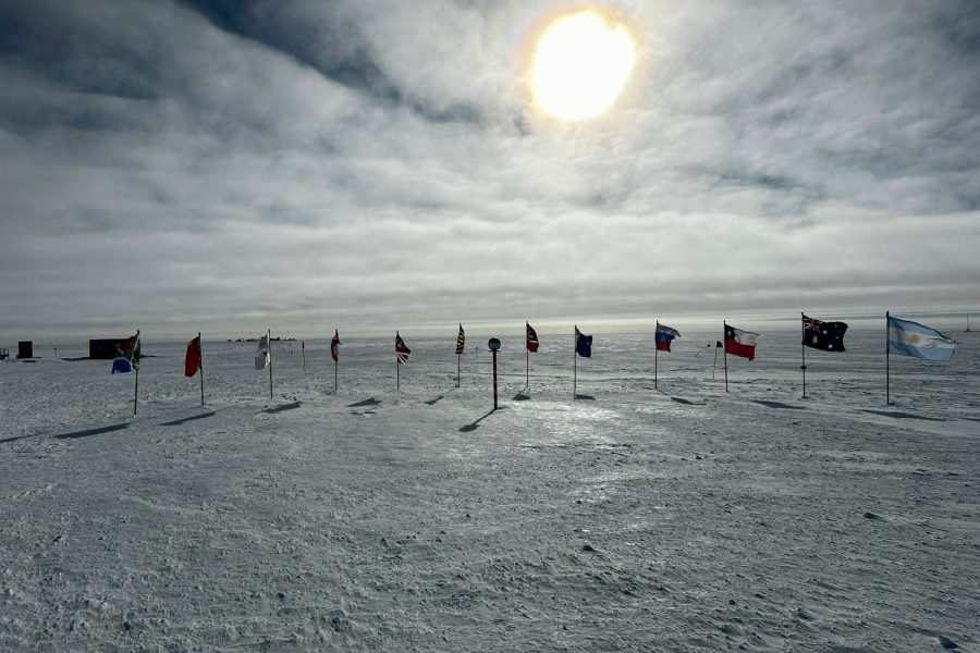 Flags from several countries stand in a row on a snowy, icy landscape under a bright sun. A few small buildings in the distance hint at an overnight trip at the South Pole, making the cold and remote scene even more captivating.