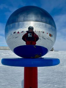 A person in red winter gear takes a photo of their reflection in a shiny metallic sphere outdoors during an overnight trip at the South Pole, with snow, a blue sky, and various flags in the background.